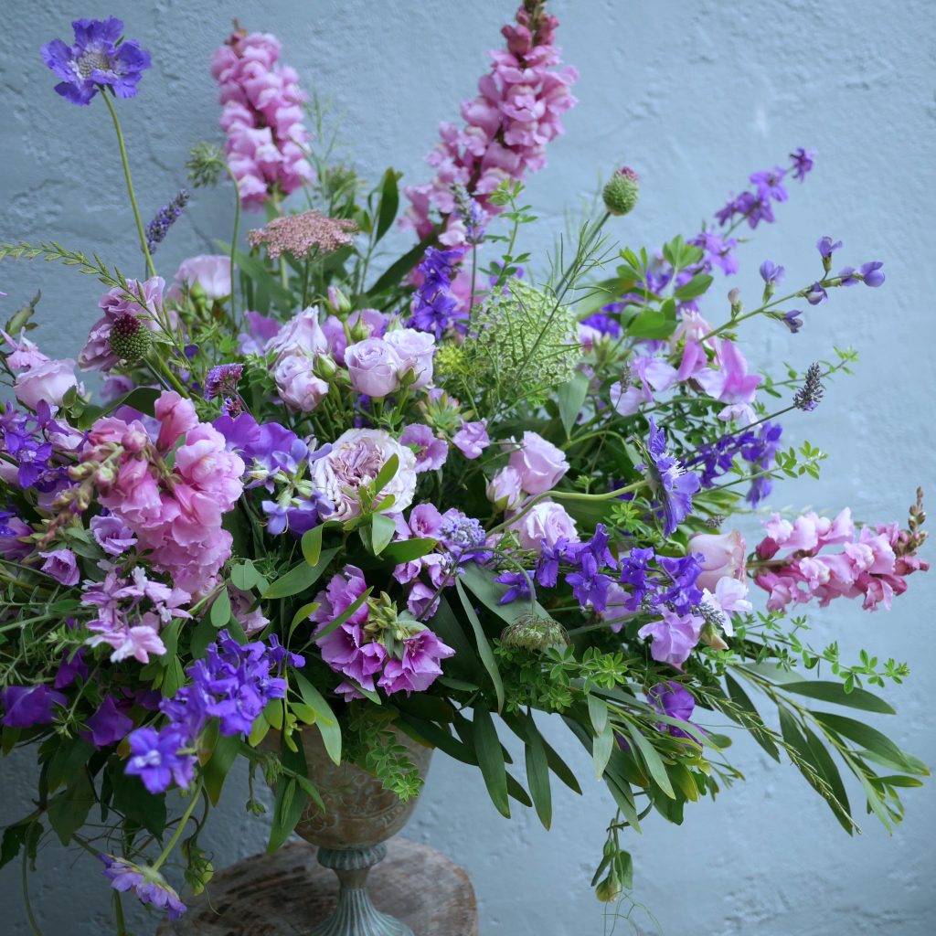 Footed vessel spilling over with lavender roses and snapdragons, purple larkspur and scabiosa, native California clarkia, Queen Anne's lace, sweet peas, baptisia, and more
